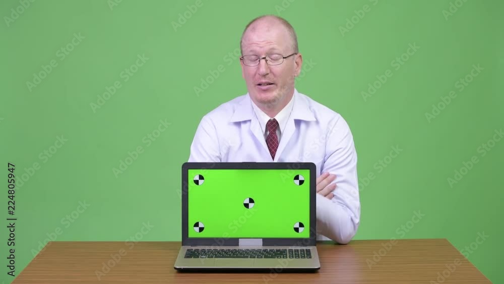 Upset mature bald man doctor showing laptop against wooden table Upset mature bald man doctor showing laptop against wooden table