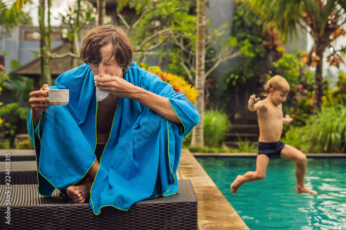 Sick man traveler. The man caught a cold on vacation, sits sad at the pool drinking tea and blows his nose into a napkin. His son is healthy and swimming in the pool. Travel insurance concept