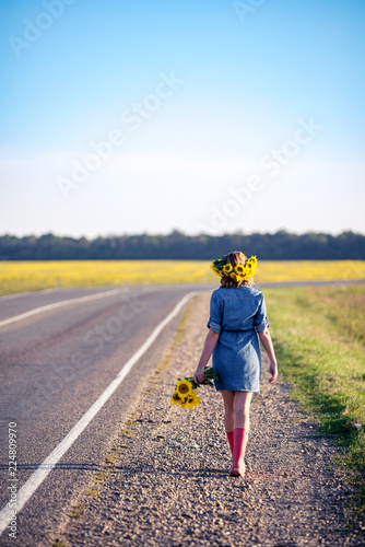 Beautiful elegant woman walking on a country road. redhaired girl in the field wreath of sunflowers and bouquet of sunflowers, dressed in a dress-shirt made of denim and red rubber boots