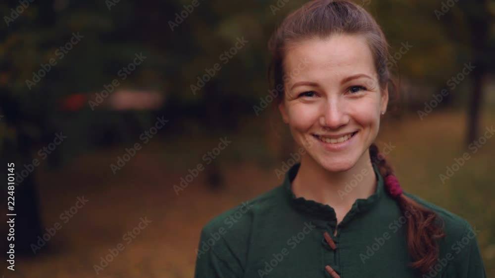 Close-up portrait of beautiful girl with braid looking at camera and smiling then laughing standing outdoors in park in autumn with trees in background. Close-up portrait of beautiful girl with braid looking at camera and smiling then laughing standing outdoors in park in autumn with trees in background.