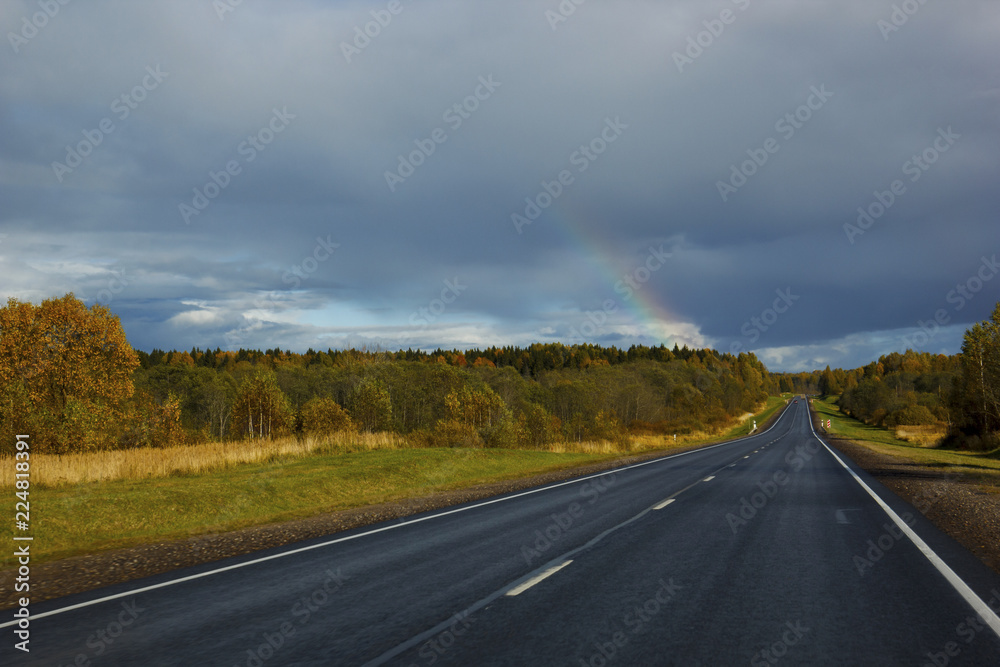 asphalt road among the fields and forests illuminated by the sun, the sky with textured clouds and clouds and a rainbow