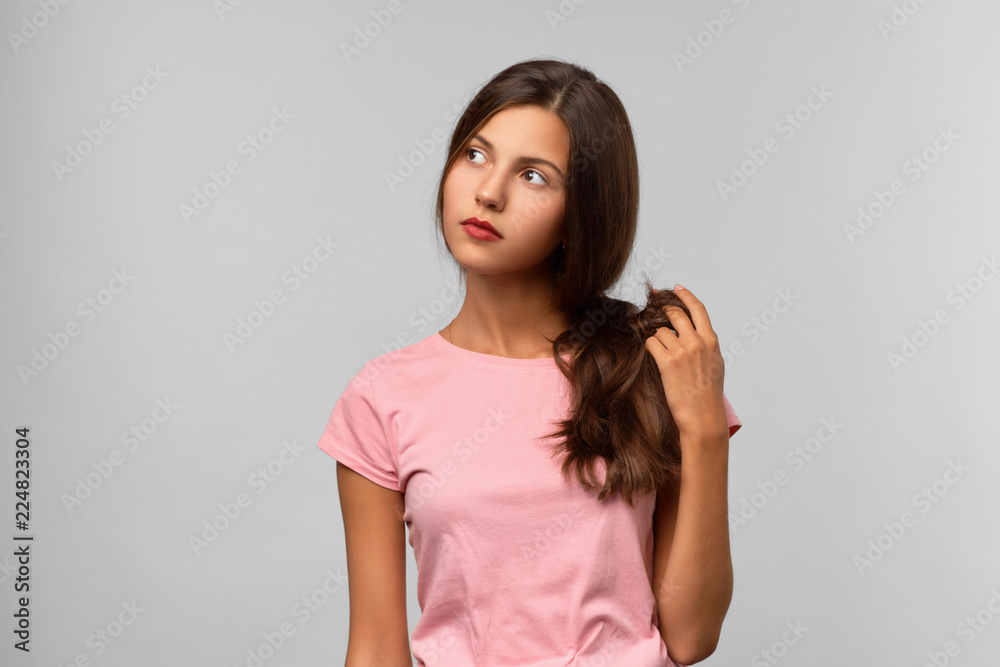 Obraz premium A beautiful young girl with long dark hair looks thoughtfully to the side and twists her hair in her hands. She faces a difficult choice and is not sure of her decision