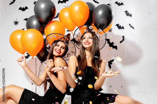 Two brunette women in witches hats smile and hold black and orange balloons. Confetti around.  Halloween