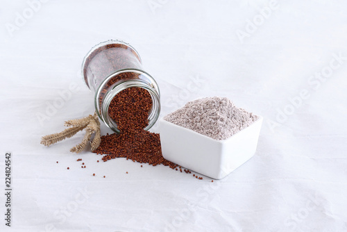 Ragi flour, which is gluten-free, in a bowl, and in the background is ragi, which is also known as finger millet, spilled from a bottle, and dried stalks.