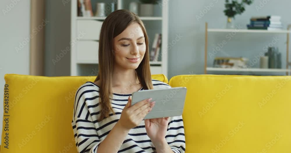 Portrait of the charming young woman in the striped blouse chatting and scrolling on the tablet device with a smile.