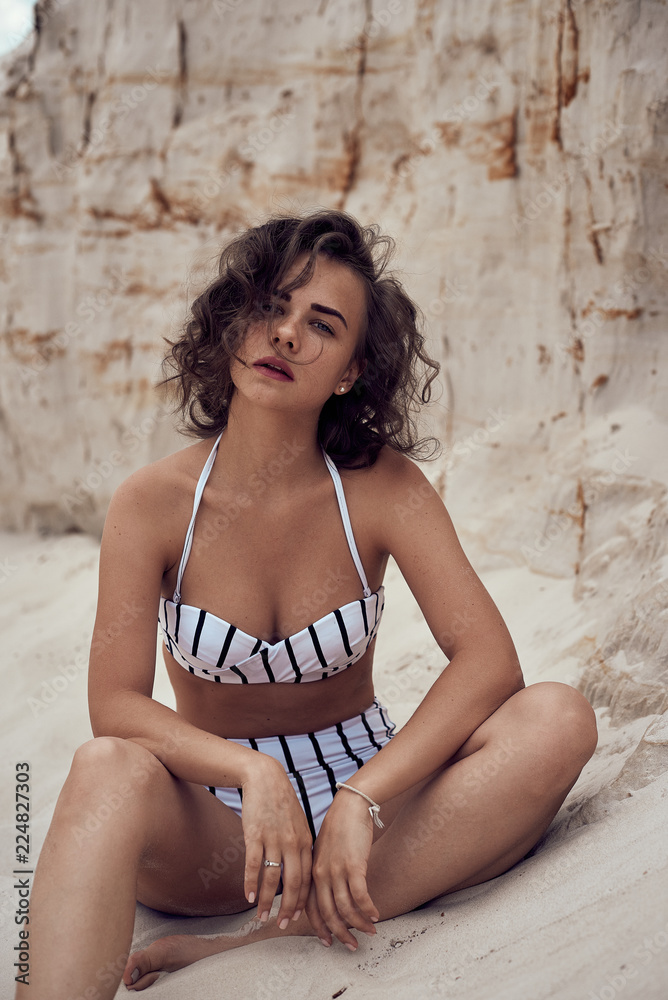 Naklejka premium portrait of beautiful caucasian sunbathed woman model with dark long hair in striped swimsuit lying on summer beach with white sand. Top view