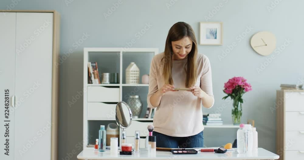 Pretty Caucasian young woman taking flat lay photo of her cosmetics which lying on the table on the smartphone. Beauty blogger in work. Pretty Caucasian young woman taking flat lay photo of her cosmetics which lying on the table on the smartphone. Beauty blogger in work.