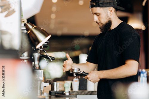 A stylish young man with beard,wearing casual clothes,cooks coffee in a coffee machine in a modern coffee shop.