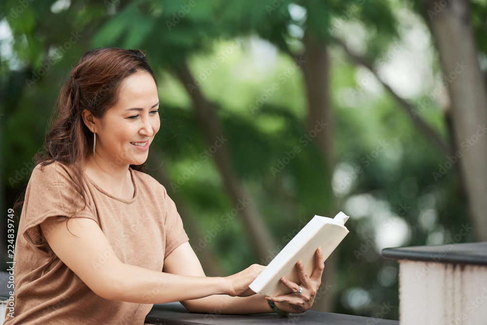 Smiling mature Vietnamese woman reading book in park Smiling mature Vietnamese woman reading book in park