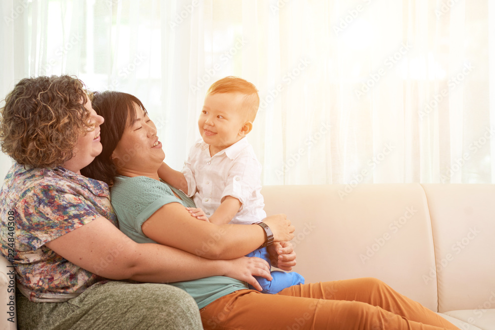 Two happy mothers playing resting on sofa with their little child on sunny day Two happy mothers playing resting on sofa with their little child on sunny day