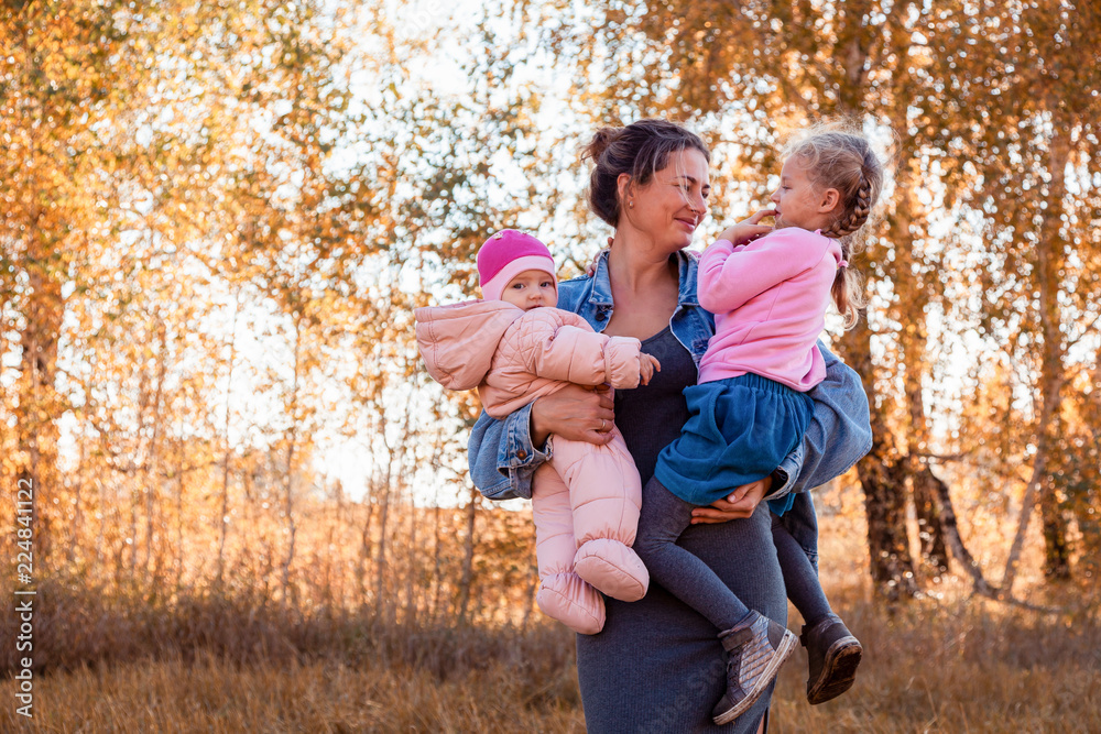 Fototapeta premium The concept of livestyle and family outdoor recreation in autumn. A young mother holding her daughters and enjoying nature, posing and talking to them on the back of a background of autumn field 