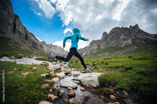 Photography Woman trail runner jumping over samll river on beautiful mountains