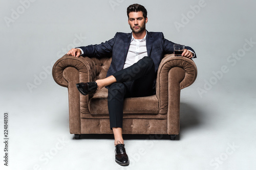 Little stylish rest. Full length of handsome young man looking at camera while sitting against white background in big chair
