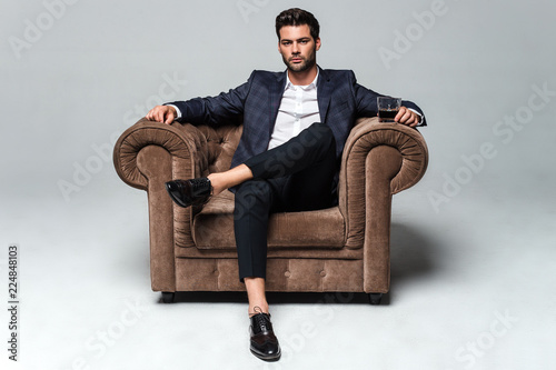 Little stylish rest. Full length of handsome young man looking at camera while sitting against white background in big chair photo