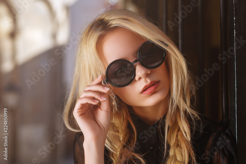 Outdoor close up portrait of young beautiful fashionable woman wearing black round sunglasses, blouse, posing in street of european city. Female fashion concept. Copy, empty space for text