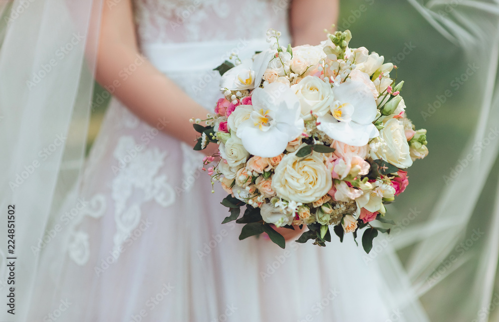 The bride in a white dress is holding a beautiful wedding bouquet in her hands. White orchids and roses. Delicate flowers.