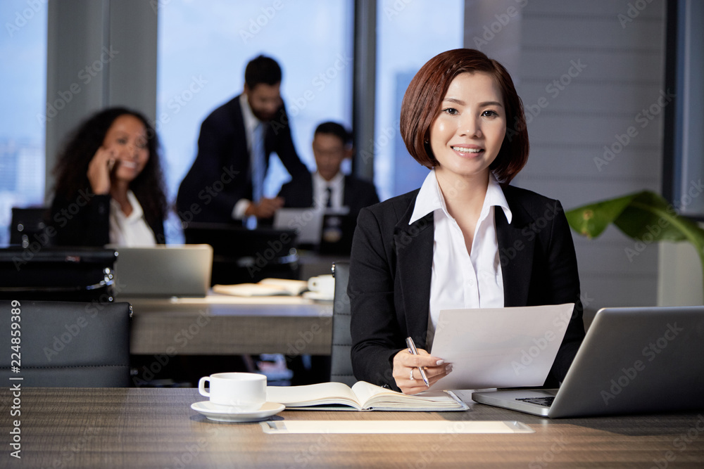 Smiling young ethnic woman with papers at table in modern office looking at camera with colleagues on background Smiling young ethnic woman with papers at table in modern office looking at camera with colleagues on background