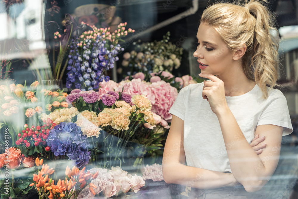 view through window of attractive pensive florist looking at bouquets in flower shop