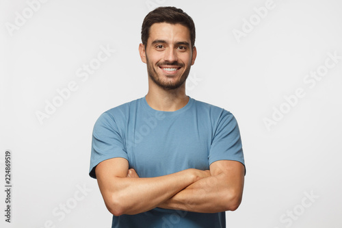 Indoor portrait of young european caucasian man isolated on gray background, standing in blue t-shirt with crossed arms, smiling and looking straight at camera