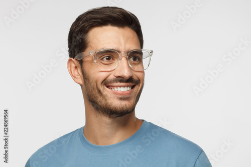 Close up portrait of smiling handsome male in blue t-shirt and transparent eyeglasses looking right, isolated on gray background © Damir Khabirov