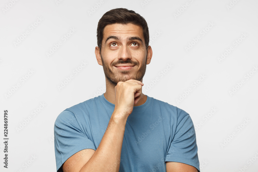 Young man in blue t-shirt with dreamy cheerful expression, thinking, looking up, isolated on gray background Young man in blue t-shirt with dreamy cheerful expression, thinking, looking up, isolated on gray background