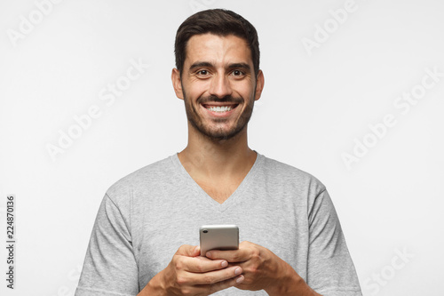 Young handsome man holding smartphone and looking at camera with smile while standing isolated on gray background