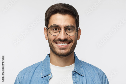 Young handsome student isolated on gray background, in blue shirt, wearing trendy round glasses, looking at camera with happy smile