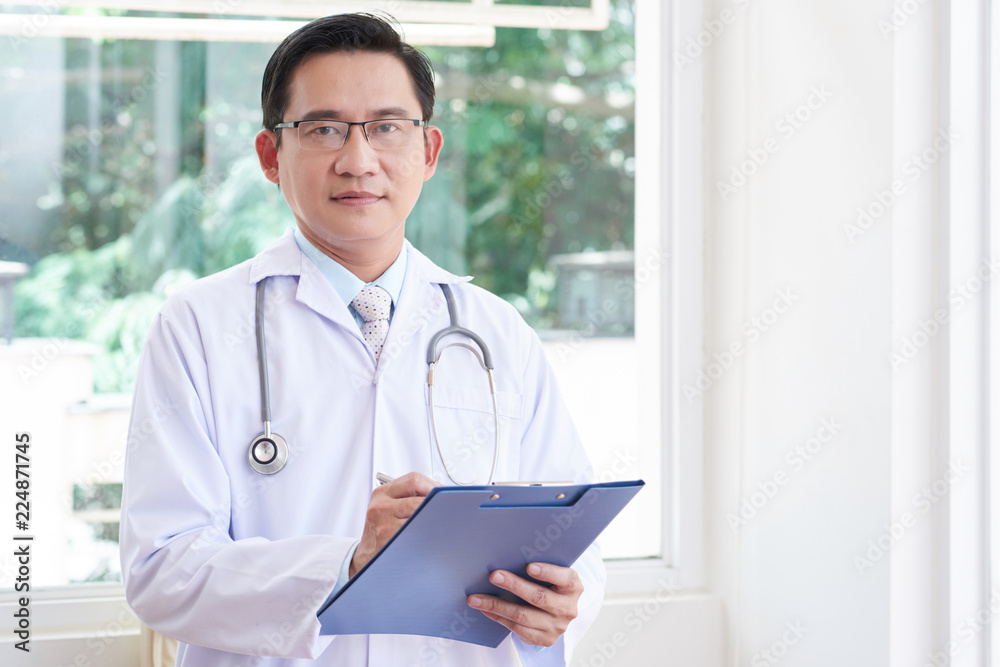 Portrait of confident male doctor in lab coat and stethoscope standing with folder at clinic Portrait of confident male doctor in lab coat and stethoscope standing with folder at clinic