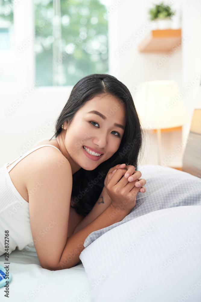 Side view of young Asian woman cuddling in cozy bed and smiling happily at camera Side view of young Asian woman cuddling in cozy bed and smiling happily at camera