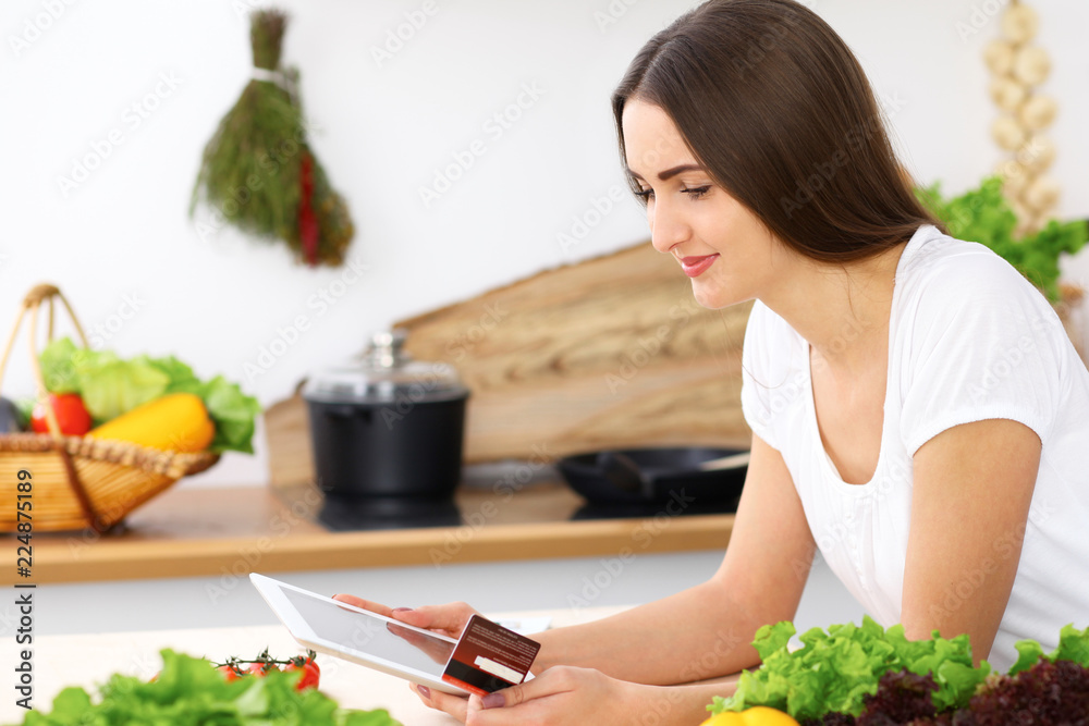 Beautiful Hispanic woman cooking while using tablet computer in kitchen or making online shopping by touchpad and credit card. Housewife found new recipe for dinner Beautiful Hispanic woman cooking while using tablet computer in kitchen or making online shopping by touchpad and credit card. Housewife found new recipe for dinner