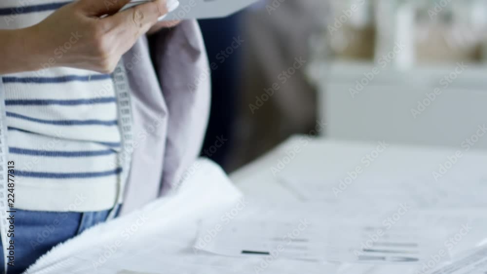 Unrecognizable young woman holding piece of fabric and color samples and using tablet computer when choosing clothing design ideas at sewing factory