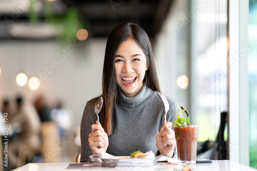 Young attractive asian woman holding fork and spoon feeling hungry, excited, happy and ready to eat healthy food looking at camera in cafe coffee shop. Charming blogger or social influencer concept.