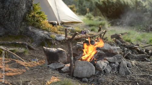 Old small kettle is heated on a bright bonfire on a green mountain meadow next to tent in the rays of rising sun - stunning morning during travel in the mountains.