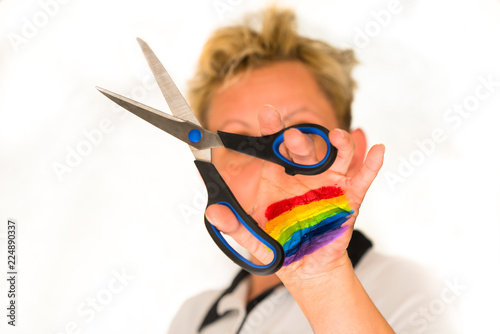 white background, at the back a blurred image of a woman with blonde hair, on the front hand holding scissors cutting the air. The hand painted in rainbow colors photo