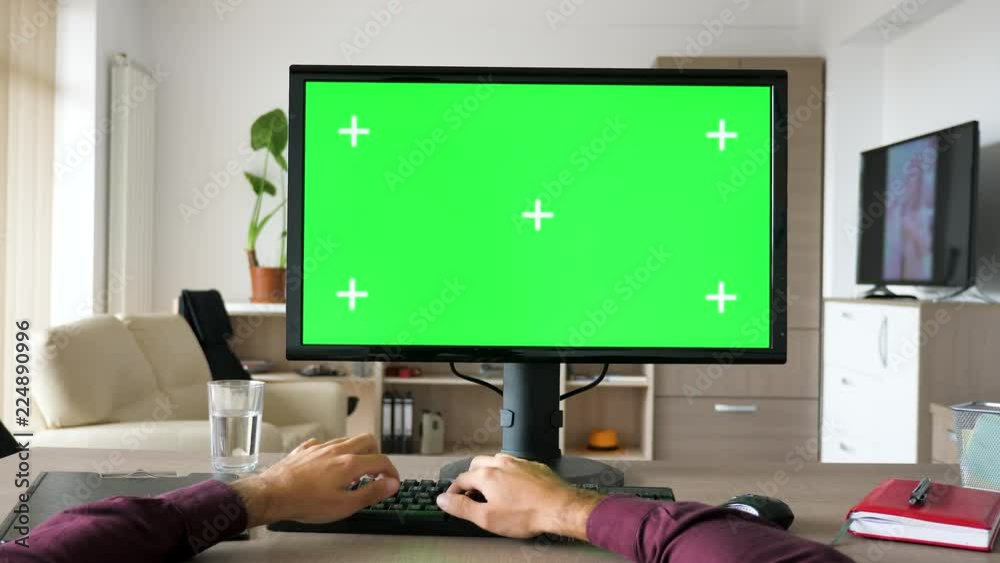 First person view - man hands typing on computer keyboard with big green screen chroma mock-up. The PC is on the desk in living room and the TV is on in the background First person view - man hands typing on computer keyboard with big green screen chroma mock-up. The PC is on the desk in living room and the TV is on in the background