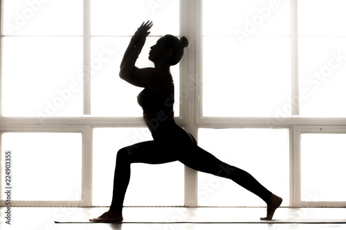 Young sporty attractive woman practicing yoga, doing Warrior one exercise, Virabhadrasana 1 pose, working out, wearing sportswear, indoor full length, at yoga studio, side view black silhouette