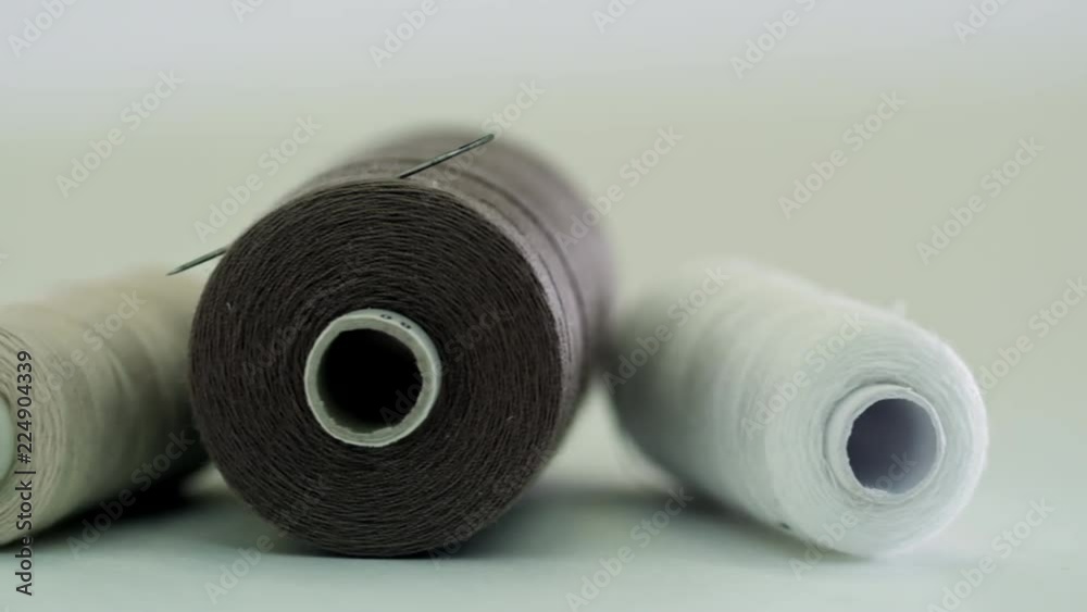 Extreme close up shot of three thread spools of white, brown and grey colors on white background, needle is stuck into one of them, tracking left Extreme close up shot of three thread spools of white, brown and grey colors on white background, needle is stuck into one of them, tracking left
