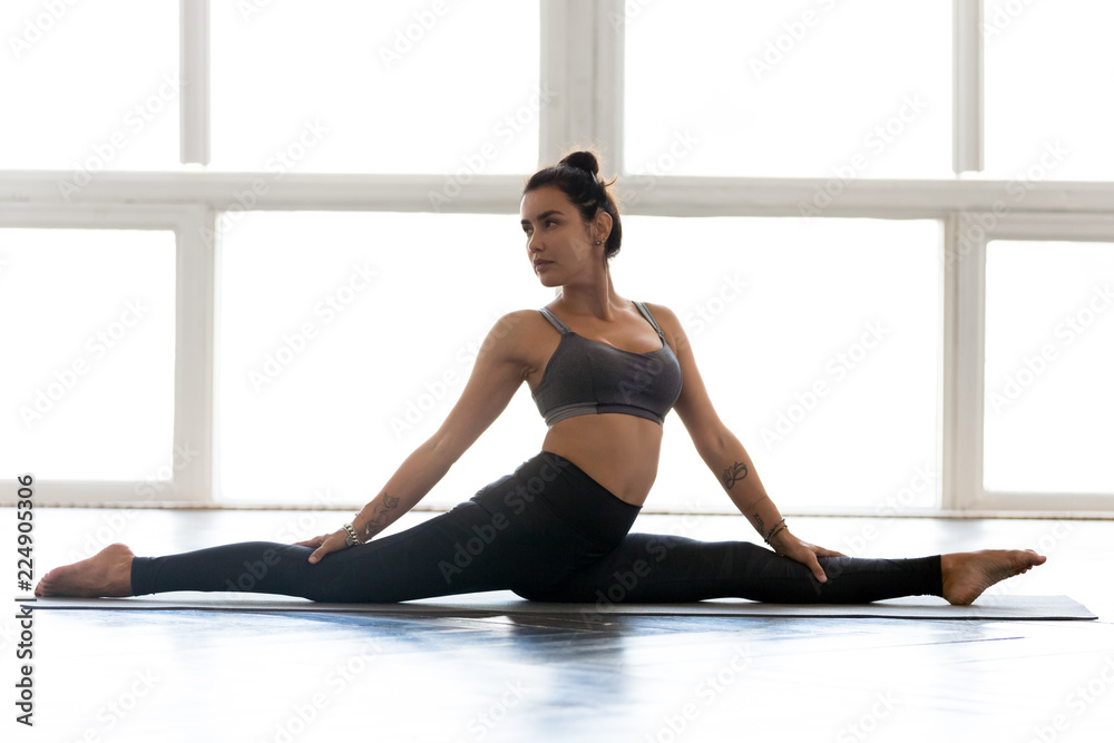 Fototapeta premium Young sporty attractive woman practicing yoga, doing Monkey God exercise, Splits, Hanumanasana pose, working out, wearing sportswear, grey pants, top, indoor full length, at yoga studio