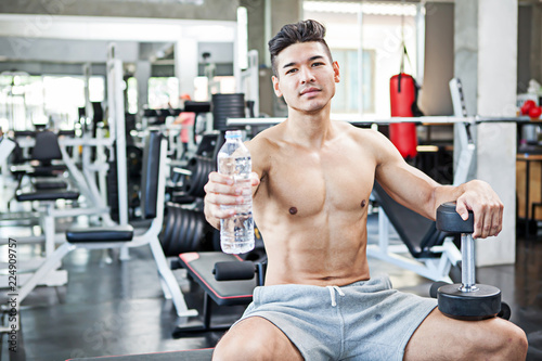 Muscular bodybuilder guy sitting on a bench show water bottle and dumbbells after exercise in fitness gym . Shirtless young sportsman resting after training at the gym