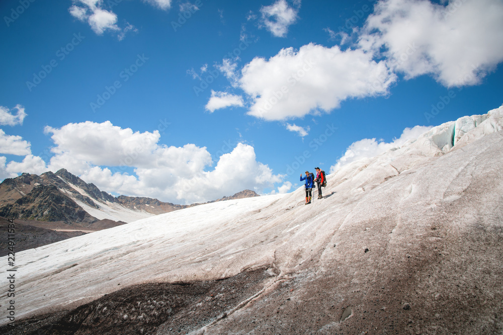 Two tourists, a man and a woman with backpacks and cats on their feet, stand on the ice against the background of the mountains of the sky and clouds. The man is showing gesture came the idea