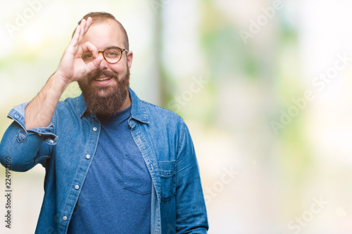 Young caucasian hipster man wearing glasses over isolated background doing ok gesture with hand smiling, eye looking through fingers with happy face.