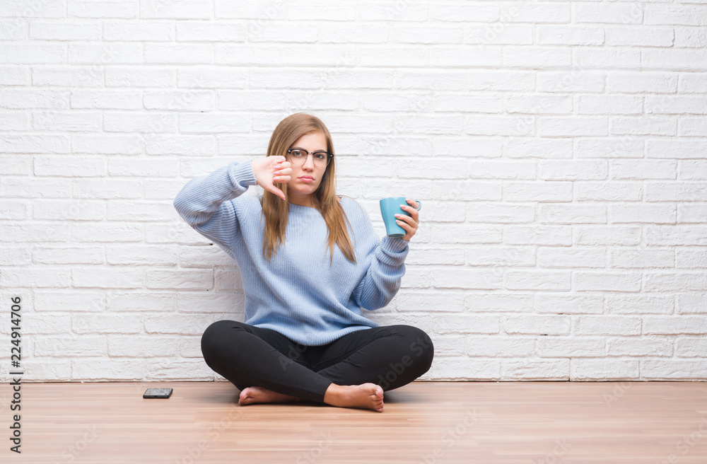 Young adult woman sitting on the floor over white brick wall driking cup of coffee with angry face, negative sign showing dislike with thumbs down, rejection concept Young adult woman sitting on the floor over white brick wall driking cup of coffee with angry face, negative sign showing dislike with thumbs down, rejection concept
