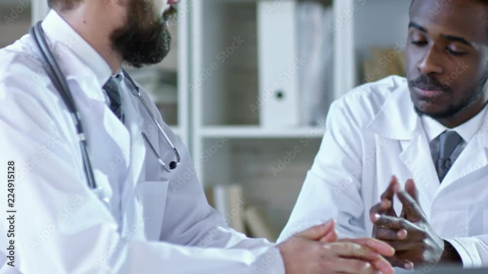 Tilt down shot of two young men in lab coats talking to each other and shaking hands when sitting at desk in office Tilt down shot of two young men in lab coats talking to each other and shaking hands when sitting at desk in office