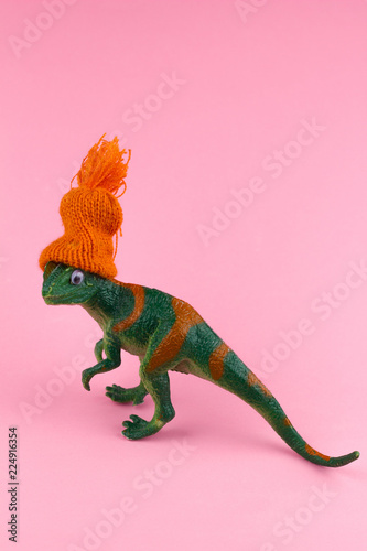 funny green dinosaur toy in little knitted orange hat  near on pastel pink background