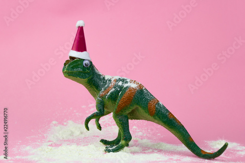 funny green dinosaur toy in little santa claus hat  and snow on pastel pink background © dvulikaia