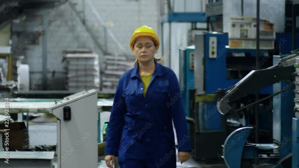 Middle-aged woman in blue workwear and yellow safety helmet walking through industrial facility Middle-aged woman in blue workwear and yellow safety helmet walking through industrial facility