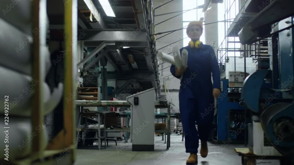 Front view of confident female engineer in workwear, hardhat and earmuffs carrying rolled blueprints along industrial facility, tracking shot Front view of confident female engineer in workwear, hardhat and earmuffs carrying rolled blueprints along industrial facility, tracking shot