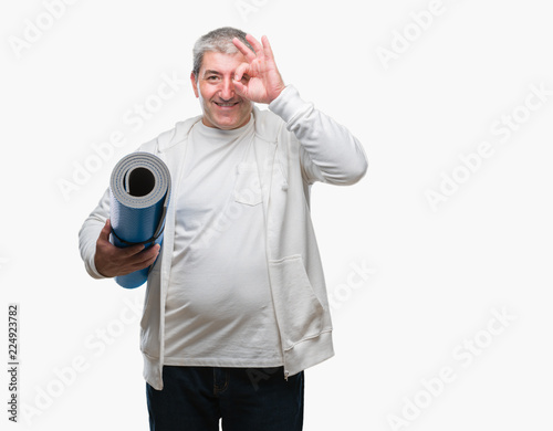 Handsome senior man holding yoga mat over isolated background with happy face smiling doing ok sign with hand on eye looking through fingers