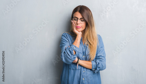 Young adult woman over grunge grey wall wearing glasses thinking looking tired and bored with depression problems with crossed arms.