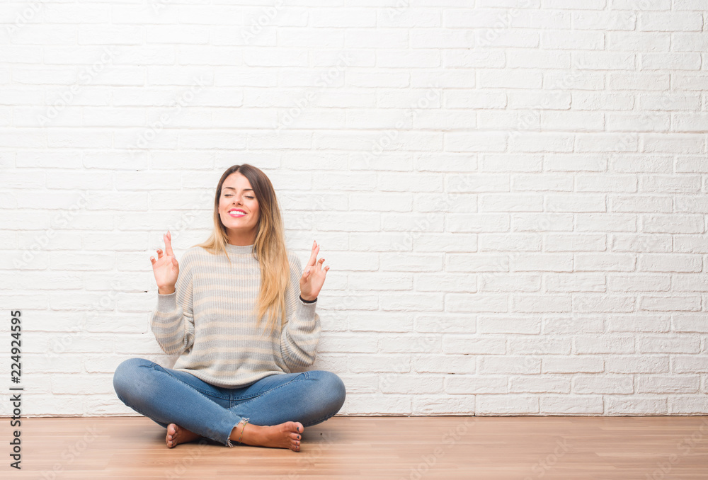Young adult woman sitting on the floor over white brick wall at home smiling crossing fingers with hope and eyes closed. Luck and superstitious concept. Young adult woman sitting on the floor over white brick wall at home smiling crossing fingers with hope and eyes closed. Luck and superstitious concept.